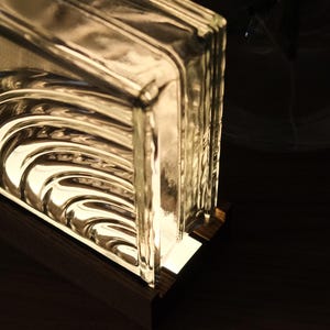 Puede incluir: Un bloque de vidrio transparente con una superficie texturizada, iluminado desde el interior. El bloque se asienta sobre una base de madera, creando un brillo cálido. El diseño presenta patrones curvos en forma de olas dentro del vidrio.