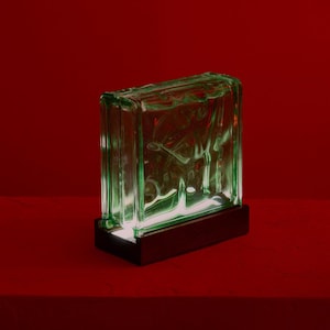 Op de afbeelding: Een groene glazen bloklamp met een houten voet. De glazen blok heeft een getextureerd, golvend patroon. De lamp is aan en geeft een warm licht.