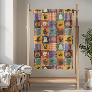 Op de afbeelding: Een rechthoekige handdoek met een patchworkpatroon van vierkanten met een Halloween-thema. Het ontwerp bevat illustraties van oranje pompoenen, zwarte katten, witte geesten, schedels, spinnenwebben, vleermuizen, heksenhoeden en ketels.