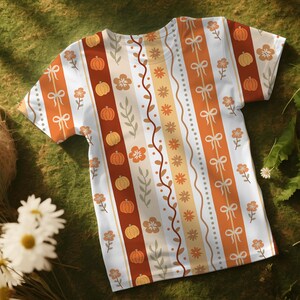 May include: White t-shirt with a vertical striped pattern featuring pumpkins, bows, flowers, and vines in shades of orange, brown, and beige. The short-sleeved shirt is made of a soft material and is suitable for casual wear.