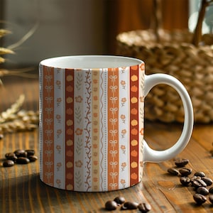 May include: White ceramic mug with a fall-themed design. The mug features vertical stripes with pumpkins, bows, and floral patterns in shades of orange, brown, and cream. The mug has a white handle and is filled with coffee beans.