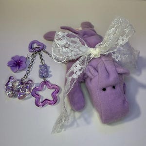 May include: Purple plush toy with a white lace bow, attached to a charm with a purple flower, a star, and a butterfly.