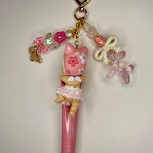 以下が含まれることがあります： ピンク色のリップグロスチューブ。ピンク色の猫のフィギュア、花のディテール、人形のようなフィギュア、金色のハート型の留め金が付いたチャームが付いています。チャームには、ピンクと白のビーズ、リボン、ピンクの風船犬も含まれています。