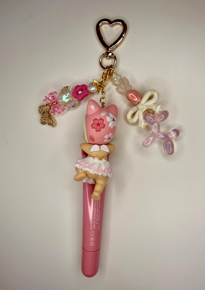 以下が含まれることがあります： ピンク色のリップグロスチューブ。ピンク色の猫のフィギュア、花のディテール、人形のようなフィギュア、金色のハート型の留め金が付いたチャームが付いています。チャームには、ピンクと白のビーズ、リボン、ピンクの風船犬も含まれています。