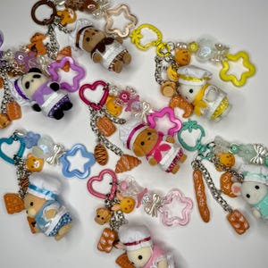 Könnte beinhalten: Eine Sammlung von Schlüsselanhängern mit Miniatur-Kochfiguren, Pandabären und verschiedenen Charms zum Thema Essen. Die Schlüsselanhänger enthalten sternförmige, herzförmige und andere geformte Akzente in einer Vielzahl von Farben mit silbernen Kettengliedern.