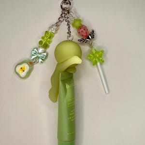 May include: A whimsical keychain featuring a green lip balm tube, a star-shaped charm, and various decorative beads. The keychain includes a green avocado charm, a strawberry charm, and a star-shaped lollipop. The lip balm tube is approximately 3 inches long.