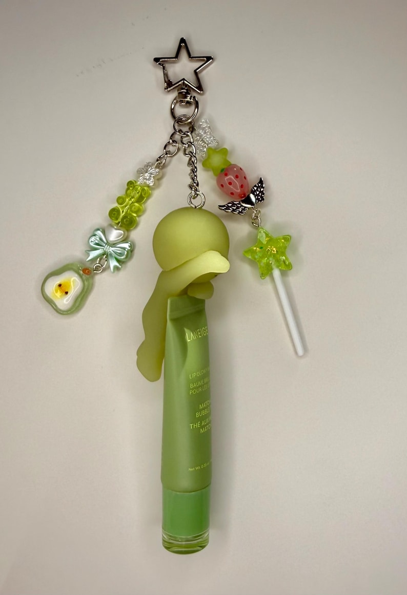 May include: A whimsical keychain featuring a green lip balm tube, a star-shaped charm, and various decorative beads. The keychain includes a green avocado charm, a strawberry charm, and a star-shaped lollipop. The lip balm tube is approximately 3 inches long.