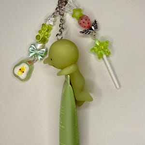 May include: A whimsical keychain featuring a silver star charm, a green figure, and various decorative beads. The keychain is attached to a green tube of product. Additional charms include a strawberry with wings, a star-shaped lollipop, and an avocado.