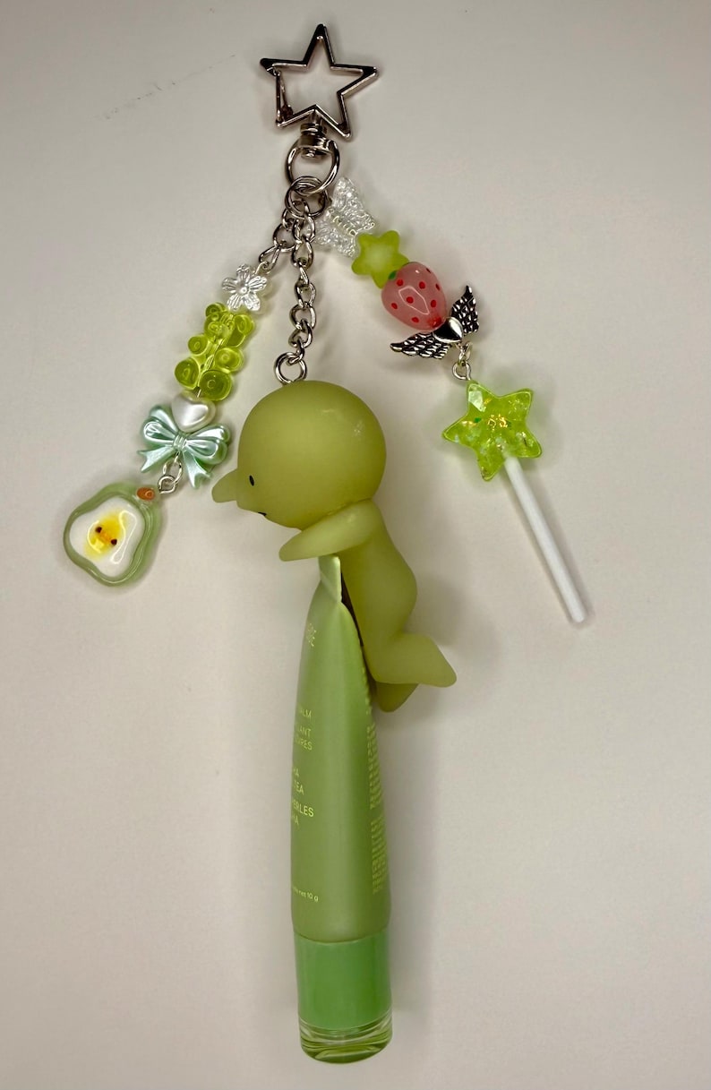 May include: A whimsical keychain featuring a silver star charm, a green figure, and various decorative beads. The keychain is attached to a green tube of product. Additional charms include a strawberry with wings, a star-shaped lollipop, and an avocado.