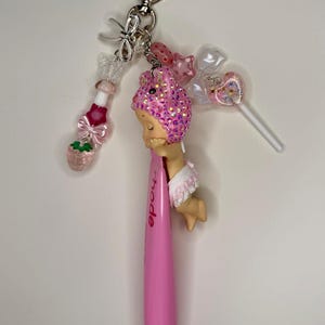 May include: A pink pen with the word "Rhode" printed on it, attached to a keychain with various charms. Charms include a strawberry, a lollipop, and a figure with a pink sequined head. The keychain has a heart-shaped clasp.