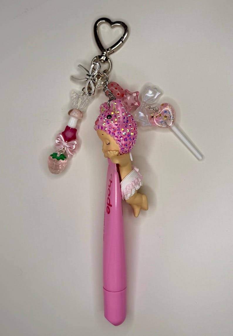 May include: A pink pen with the word "Rhode" printed on it, attached to a keychain with various charms. Charms include a strawberry, a lollipop, and a figure with a pink sequined head. The keychain has a heart-shaped clasp.