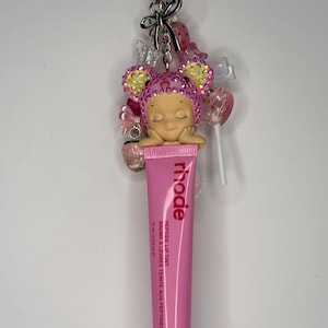 May include: A pink lip tint tube with the brand name "rhode" and the text "Peptide Lip Tint" is attached to a keychain. The keychain also features a decorative charm with a sleeping doll, bows, and candy-like embellishments.
