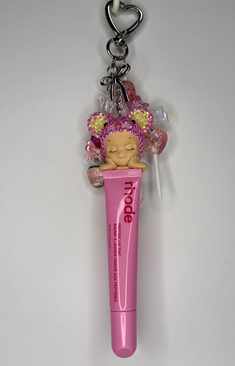 May include: A pink lip tint tube with the brand name "rhode" and the text "Peptide Lip Tint" is attached to a keychain. The keychain also features a decorative charm with a sleeping doll, bows, and candy-like embellishments.