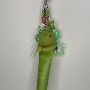 May include: A green Laneige lip balm tube is attached to a keychain with a green cartoon character, star-shaped charms, and beads. The lip balm is labeled "Matcha Bubble Tea".