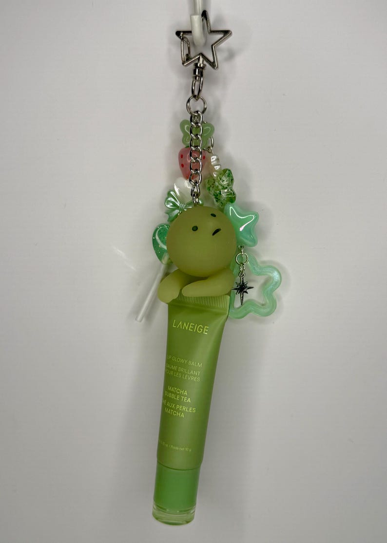 May include: A green Laneige lip balm tube is attached to a keychain with a green cartoon character, star-shaped charms, and beads. The lip balm is labeled "Matcha Bubble Tea".
