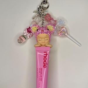 May include: A pink lip tint tube with the word "rhode" printed on it, attached to a keychain with a heart-shaped clasp. The keychain features a charm of a sleeping figure wearing a pink mouse ear hat, and various decorative beads and charms.