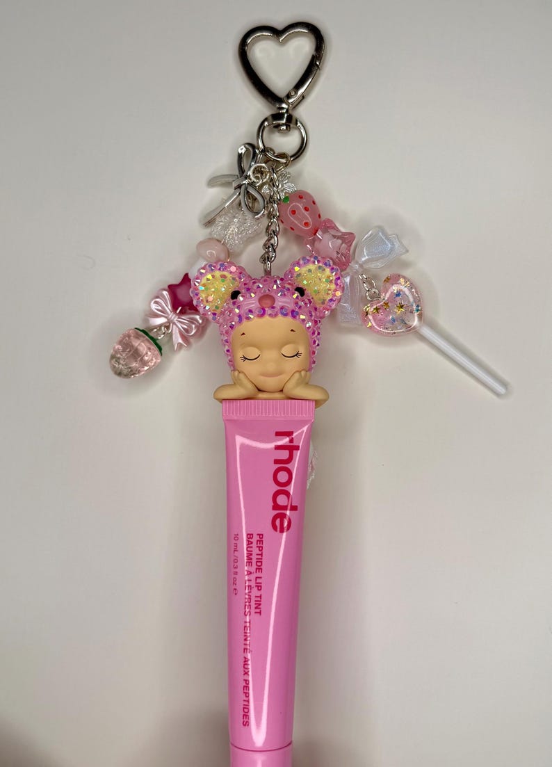 May include: A pink lip tint tube with the word "rhode" printed on it, attached to a keychain with a heart-shaped clasp. The keychain features a charm of a sleeping figure wearing a pink mouse ear hat, and various decorative beads and charms.