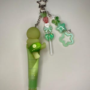 May include: A green lip butter tube with a Smiski charm and a star-shaped keychain. The keychain features a chain with green beads, a strawberry, a bow, a heart-shaped lollipop, and green star charms.
