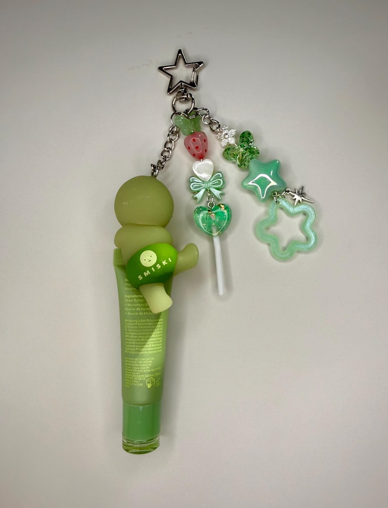 May include: A green lip butter tube with a Smiski charm and a star-shaped keychain. The keychain features a chain with green beads, a strawberry, a bow, a heart-shaped lollipop, and green star charms.