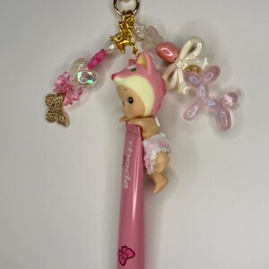 以下が含まれることがあります： ピンク色のキーホルダー。ピンクのキツネの帽子とオムツをつけた小さな人形が、ピンク色の口紅チューブに付いています。キーホルダーには、金色のハート型の留め金、装飾ビーズ、チャームが含まれています。