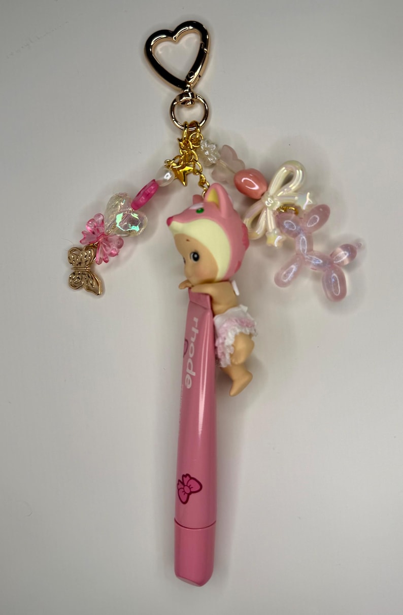 以下が含まれることがあります： ピンク色のキーホルダー。ピンクのキツネの帽子とオムツをつけた小さな人形が、ピンク色の口紅チューブに付いています。キーホルダーには、金色のハート型の留め金、装飾ビーズ、チャームが含まれています。