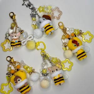 May include: Five keychains featuring small animal figures dressed in yellow and black striped bee costumes. Each keychain includes a metal clasp, a star-shaped charm, various yellow and clear beads, small butterfly charms, and a fluffy white pom-pom.