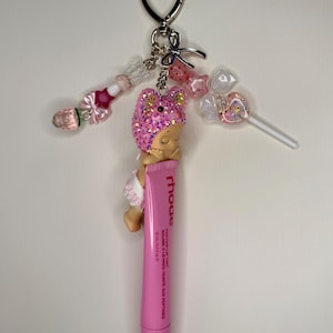 May include: A pink lip tint tube with a sleeping baby charm, a heart-shaped keychain, and beaded accents. The charm has a pink glittery hat. The lip tint tube has the brand name "rhoe" and the text "peptide lip tint".