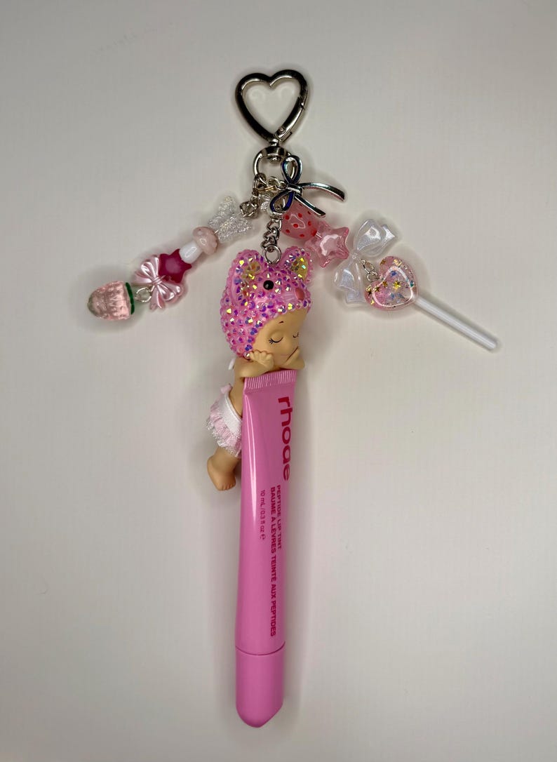 May include: A pink lip tint tube with a sleeping baby charm, a heart-shaped keychain, and beaded accents. The charm has a pink glittery hat. The lip tint tube has the brand name "rhoe" and the text "peptide lip tint".