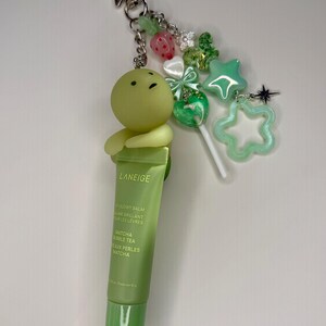 May include: A green LANEIGE lip balm keychain with a cartoon character, star, and other charms. The lip balm tube is green with the text "Matcha Bubble Tea". The keychain is attached to a silver star-shaped clasp.
