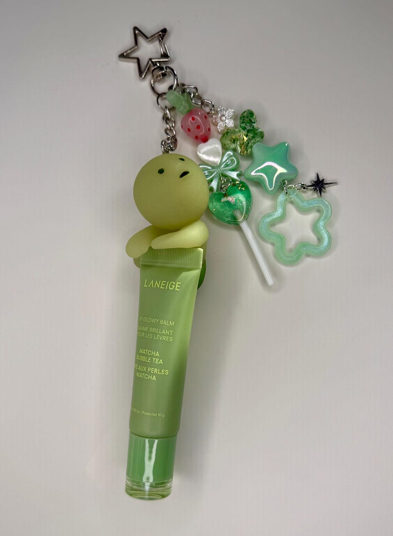 May include: A green LANEIGE lip balm keychain with a cartoon character, star, and other charms. The lip balm tube is green with the text "Matcha Bubble Tea". The keychain is attached to a silver star-shaped clasp.