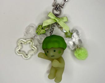 Puede incluir: Un llavero caprichoso con una pequeña figura verde claro con una gorra en forma de hongo. El llavero incluye un cierre en forma de estrella, un lazo verde, dijes transparentes de mariposa y estrella y un pompón verde esponjoso.