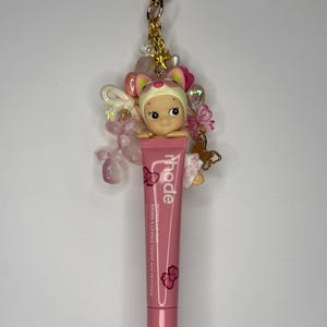 以下が含まれることがあります： ピンク色のRhodeリップティントチューブと装飾的なチャーム。チャームには小さな人形、ピンクの花、金色のハート型のキーホルダーが付いています。チューブには「Rhode」と「Baume &agrave; l&egrave;vres teint&eacute; aux peptides」の文字があります。