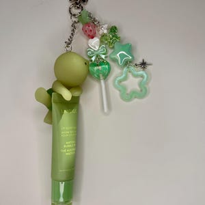 May include: A green lip balm tube with a turtle charm, star, and bead accents. The tube reads "LANEIGE LIP GLOWY BAUME". The keychain includes a lollipop charm and a star-shaped charm. The overall theme is a cute, whimsical accessory.