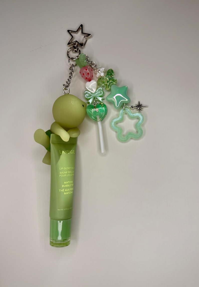May include: A green lip balm tube with a turtle charm, star, and bead accents. The tube reads "LANEIGE LIP GLOWY BAUME". The keychain includes a lollipop charm and a star-shaped charm. The overall theme is a cute, whimsical accessory.