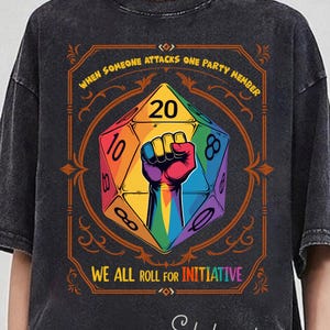 May include: Dark gray t-shirt with a colorful graphic featuring a rainbow-colored fist inside a D20 die. Text reads "WHEN SOMEONE ATTACKS ONE PARTY MEMBER" and "WE ALL ROLL FOR INITIATIVE". The shirt has a vintage, distressed look.