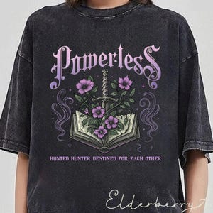 May include: Black t-shirt with a graphic of a book with a sword and flowers. The text "Powerless" is above the graphic and the text "Hunted Hunter Destined for Each Other" is below the graphic.