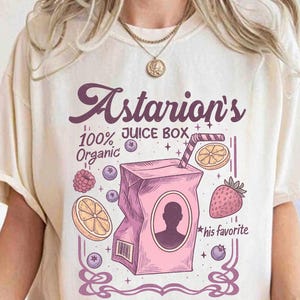 Könnte beinhalten: Cremefarbenes T-Shirt mit einem rosa Saftbox-Motiv und dem Text "Astarion's 100% Juice Box Organic". Das Design beinhaltet Obstillustrationen und den Satz "his favorite".