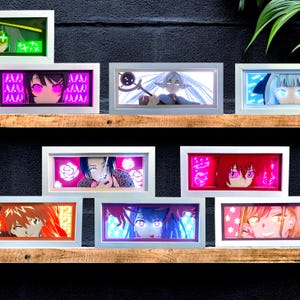 May include: A collection of framed anime-style art pieces, each featuring a different character's face with glowing accents. The frames are white, and the artwork is displayed on wooden shelves against a dark background. The art pieces have a variety of colors.