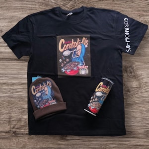 May include: A black t-shirt with a cartoon image of a man eating cereal. The cereal box says "Cornholio's" and the man is wearing a blue robe. The t-shirt also has the words "Cornholio's" printed on the sleeve. There is also a brown beanie with the same image and a can of the cereal.