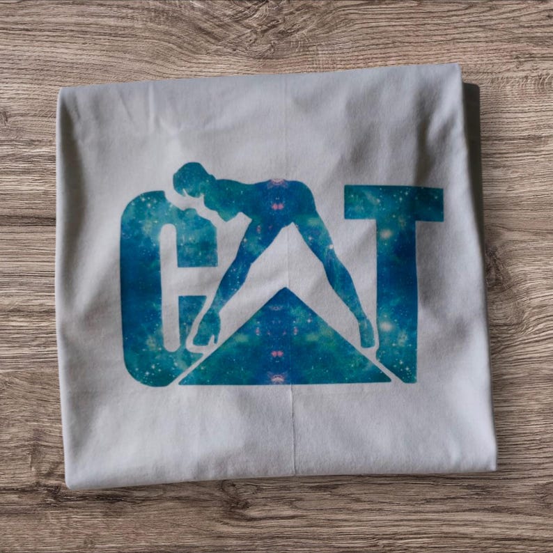 Puede incluir: Camiseta blanca con una impresi&oacute;n de galaxia azul y verde de la palabra "CAT" con una silueta de una persona en una pose sugerente.
