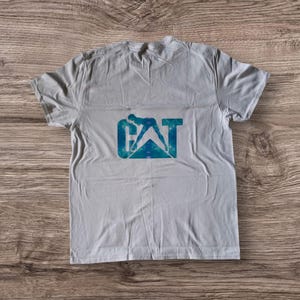 Puede incluir: Una camiseta blanca con un dise&ntilde;o tie-dye azul y verde del logotipo de CAT. El logotipo presenta una silueta de una persona operando una m&aacute;quina pesada.