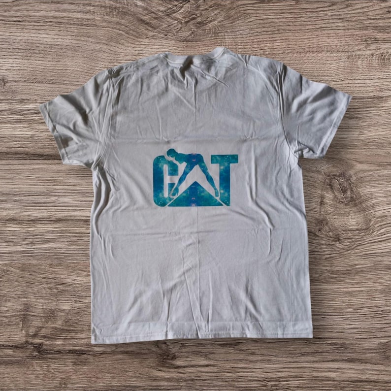 Puede incluir: Una camiseta blanca con un dise&ntilde;o tie-dye azul y verde del logotipo de CAT. El logotipo presenta una silueta de una persona operando una m&aacute;quina pesada.