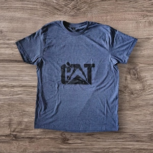 K&ouml;nnte beinhalten: Ein blaues T-Shirt mit einem schwarzen CAT-Logo, das die Silhouette einer Person zeigt, die ein Baumaschinen bedient.