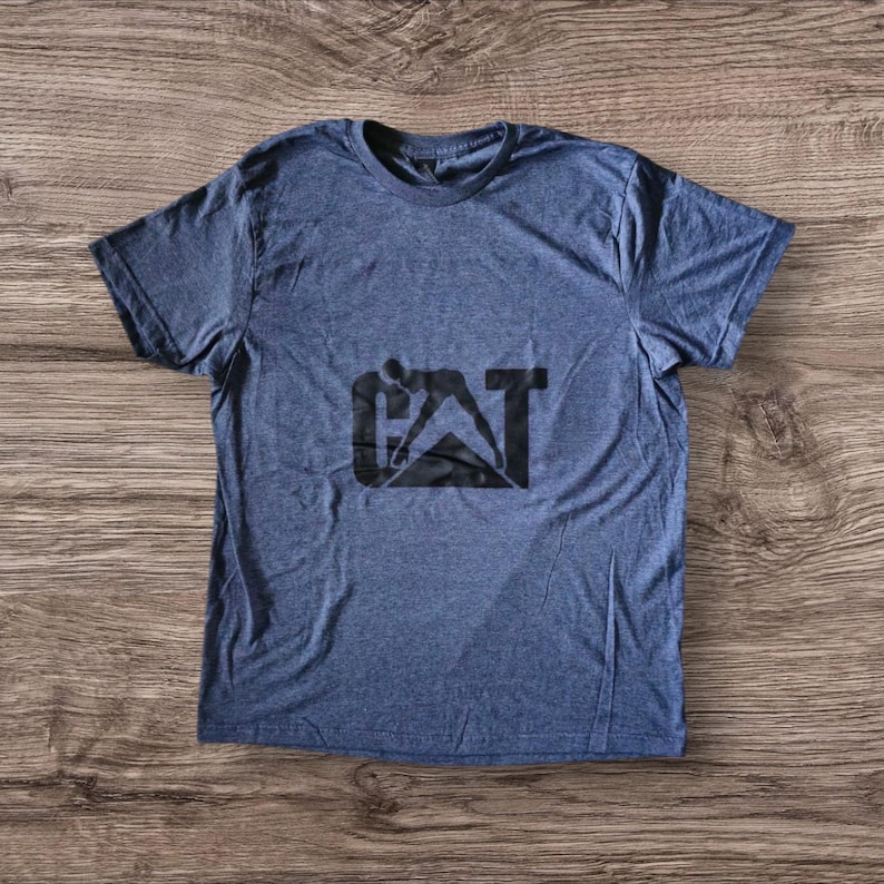 K&ouml;nnte beinhalten: Ein blaues T-Shirt mit einem schwarzen CAT-Logo, das die Silhouette einer Person zeigt, die ein Baumaschinen bedient.