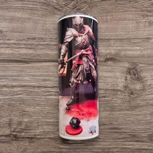 Puede incluir: Un vaso de acero inoxidable blanco con una imagen en negro y rojo de un personaje de fantasía con armadura sosteniendo una espada. El personaje está de pie sobre un tablero de ajedrez rojo y blanco con un dado negro y blanco en un círculo rojo.
