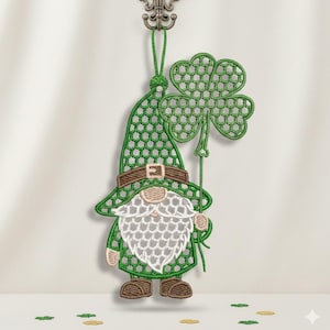 May include: A green embroidered gnome holding a shamrock. The gnome has a white beard, brown shoes, and a green hat with a brown band. The shamrock is also green. The design is hanging from a decorative hook.