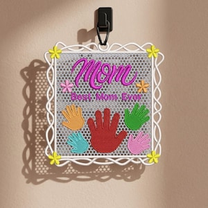 May include: Embroidered square wall hanging with the word "Mom" in pink script. The design includes colorful handprints and the text "Best Mom Ever." The frame is white with yellow flower accents.