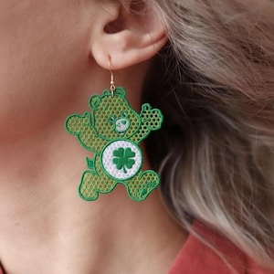 May include: Green bear-shaped earrings with a four-leaf clover design. The earrings are made of a mesh-like material and have gold-colored hooks. The bear has a white belly with a green clover. The earrings are a fun accessory.