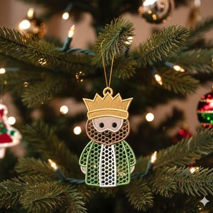Può includere: Ornamento natalizio ricamato raffigurante un re con corona d'oro, barba e veste verde. L'ornamento è appeso a un filo d'oro su uno sfondo sfocato di un albero di Natale con luci e altre decorazioni.