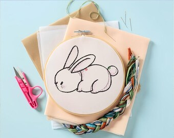 Puede incluir: Un aro de bordado blanco con un dibujo de línea simple de un conejo blanco. El aro está rodeado de materiales de artesanía, incluyendo hilo de bordar, tijeras y agujas.